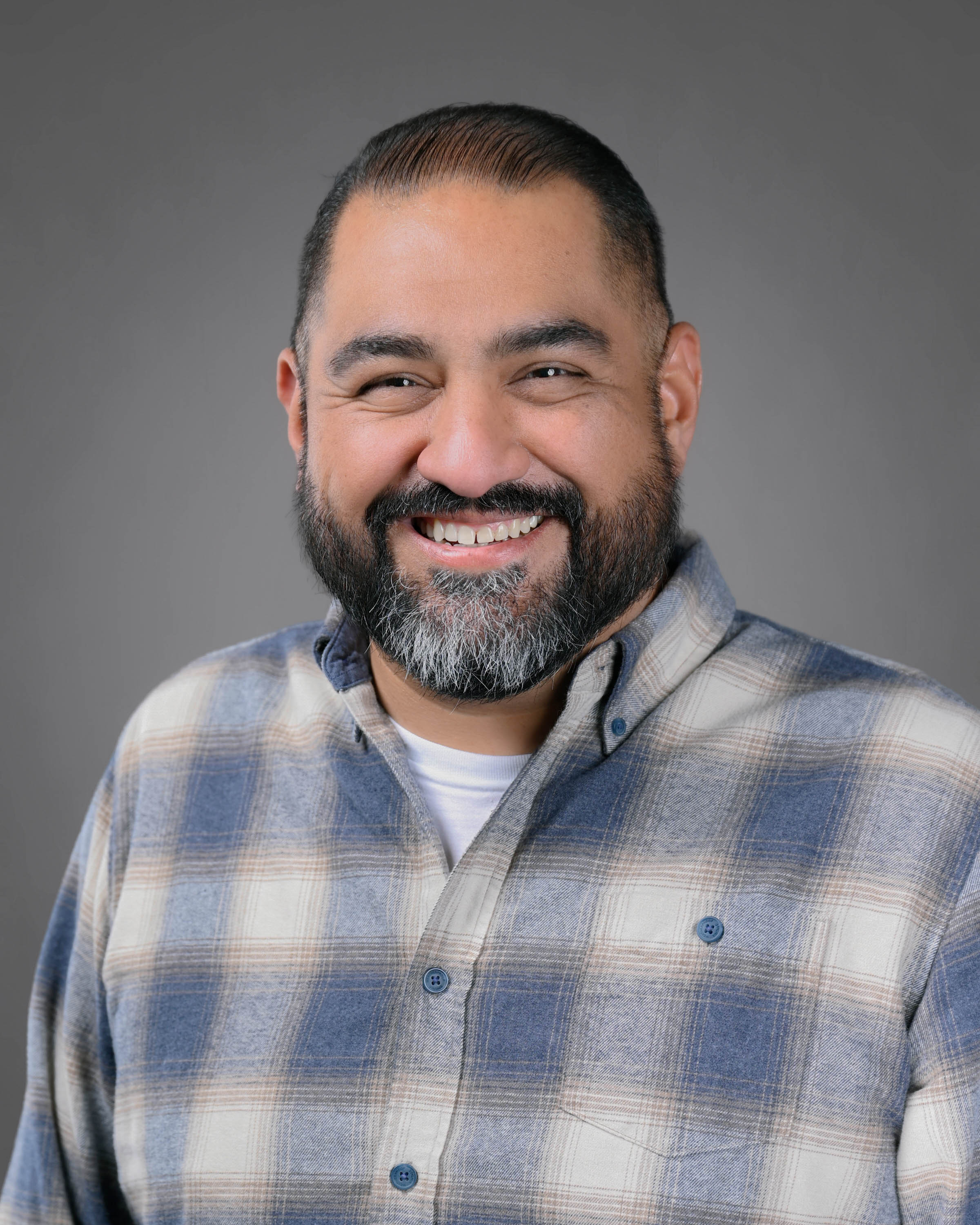 Dark haired man with a beard wearing a blue plaid shirt.